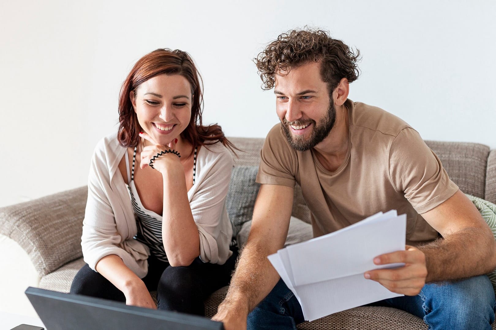Photo of smiling couple on couch looking at computer with paperwork in hand
