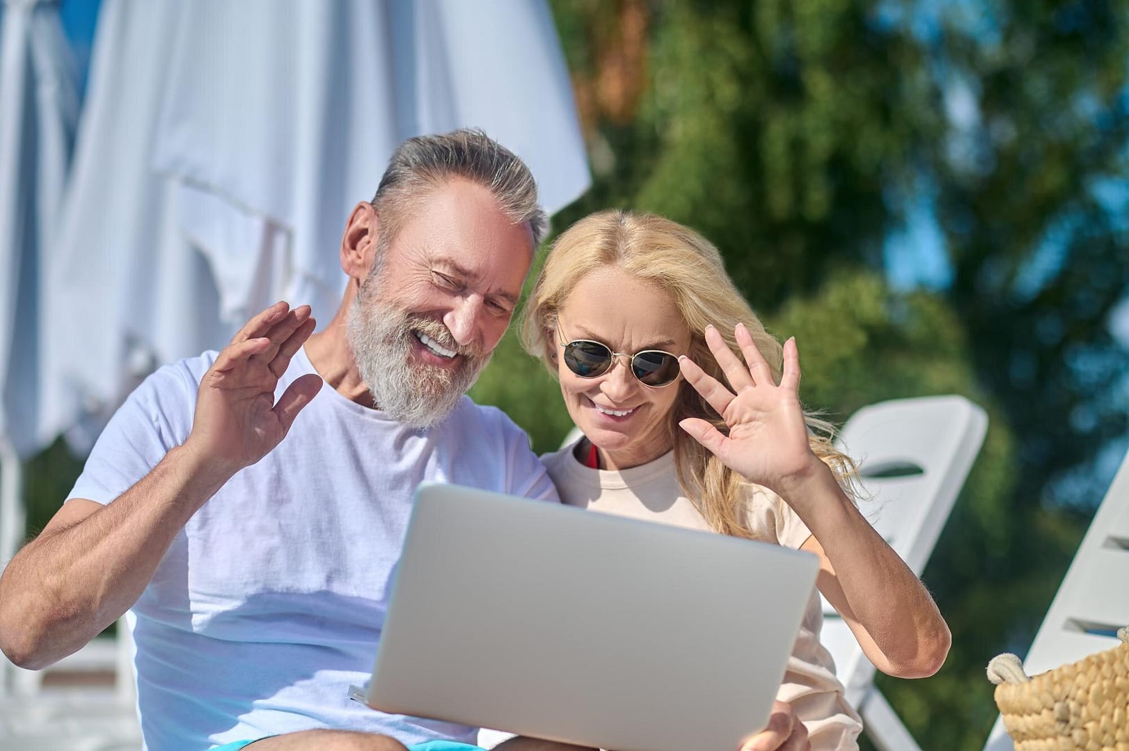 Mature couple having a video call and waving their hands smiling on beach chairs